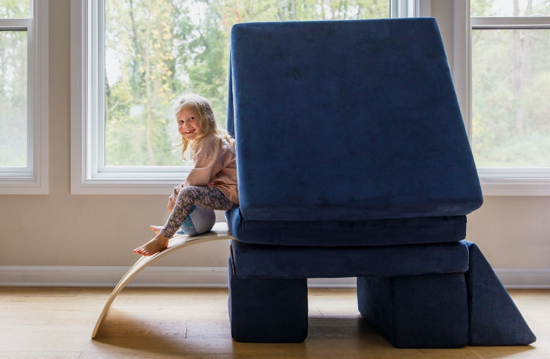 A joyful young girl sitting on a curved wooden indoor slide next to a large blue foam block chair, smiling near a window.