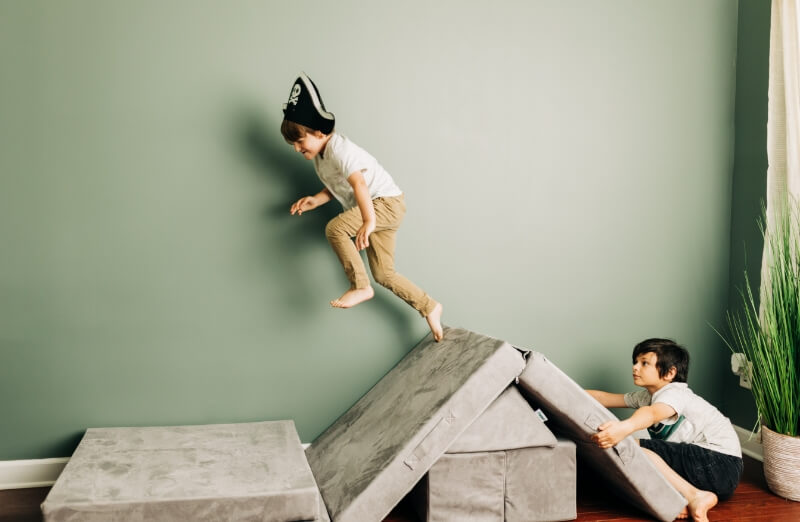 Two children play indoors, one climbing on grey foam blocks while the other watches. 500-3201-GRY