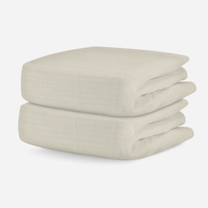 A stack of two soft, pale beige muslin swaddle blankets on a white background. skip