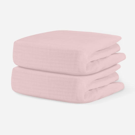 Light pink, breathable cotton crib sheets stacked neatly on a plain background. skip
