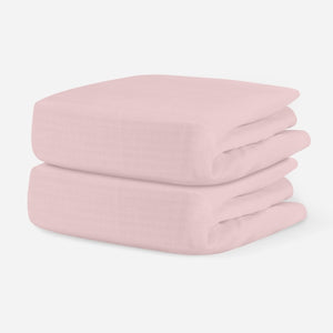 Light pink, breathable cotton crib sheets stacked neatly on a plain background. skip