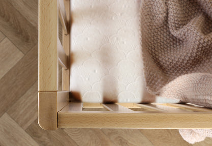 Close-up of a light wood crib with a breathable crib mattress adorned with a geometric pattern, and a soft tan knit blanket draped over the side.
